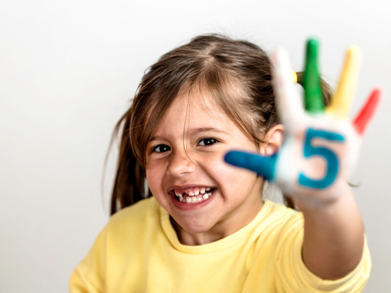 Smiling little girl with missing tooth, holding up hand with number 5 painted on palm and fingers painted different colors.