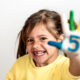 Smiling little girl with missing tooth, holding up hand with number 5 painted on palm and fingers painted different colors.