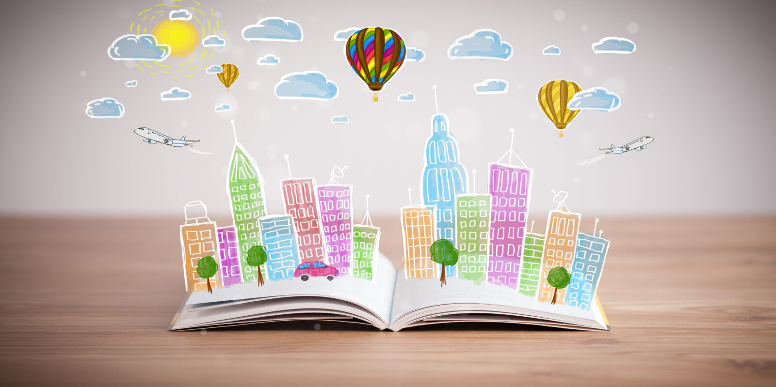 Open book with imaginative items coming out of the pages, such as colorful buildings, hot air balloons, airplanes, and clouds.