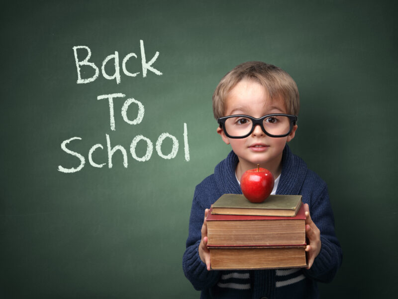 child standing in front of chalkboard that has "back to school" written on it, holding a stack of books with an apple on top