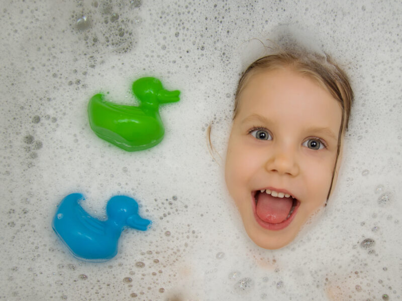 Childs happy face showing through a pile of bubbles in the bathtub next to a green and a blue rubber duck.