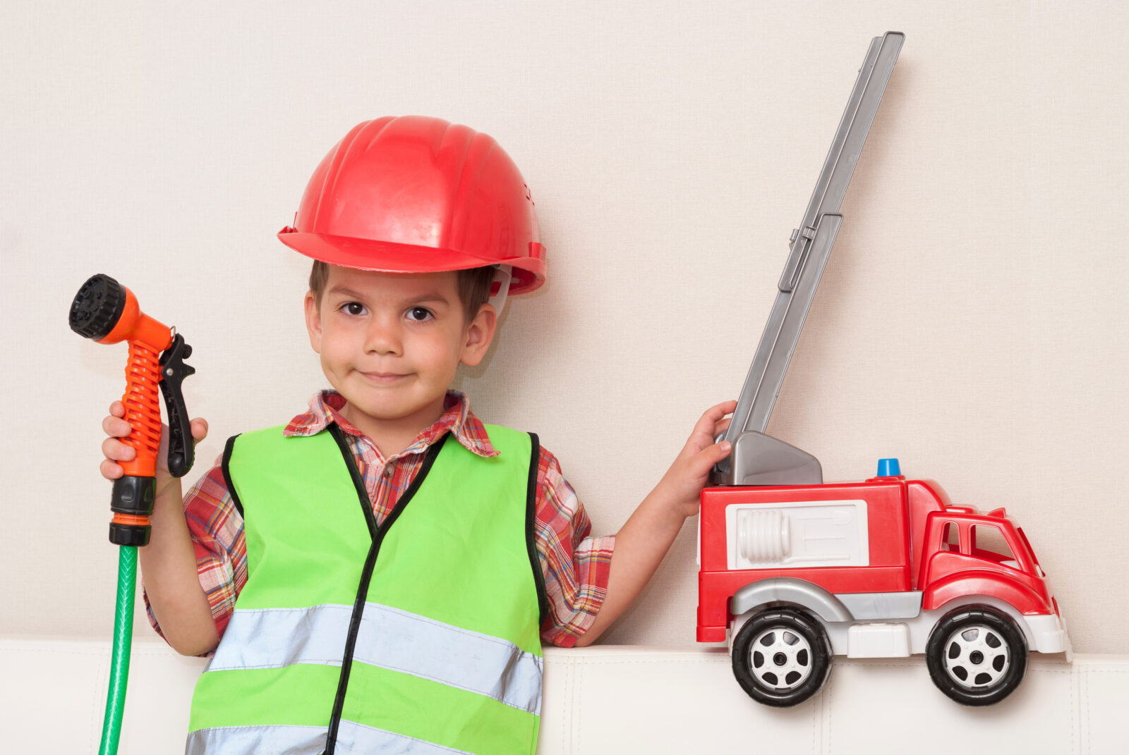 Little boy dressed in safety gear holding a toy firetruck and water hose.