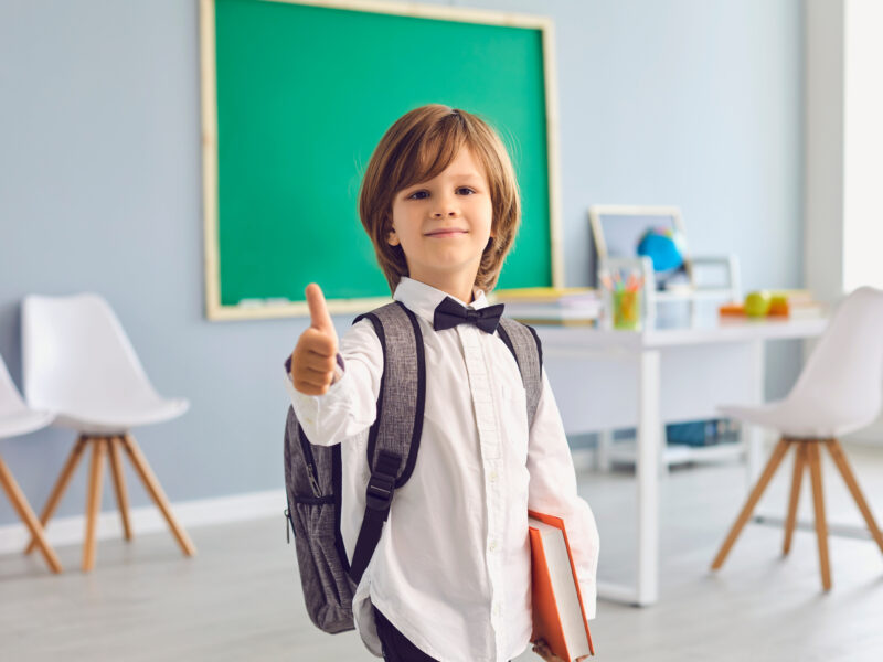 Little boy in a classroom with his backpack on, holding a book and giving a thumbs up